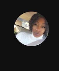 A woman with dark curly hair wearing a white turtleneck sweater, sitting in a car.