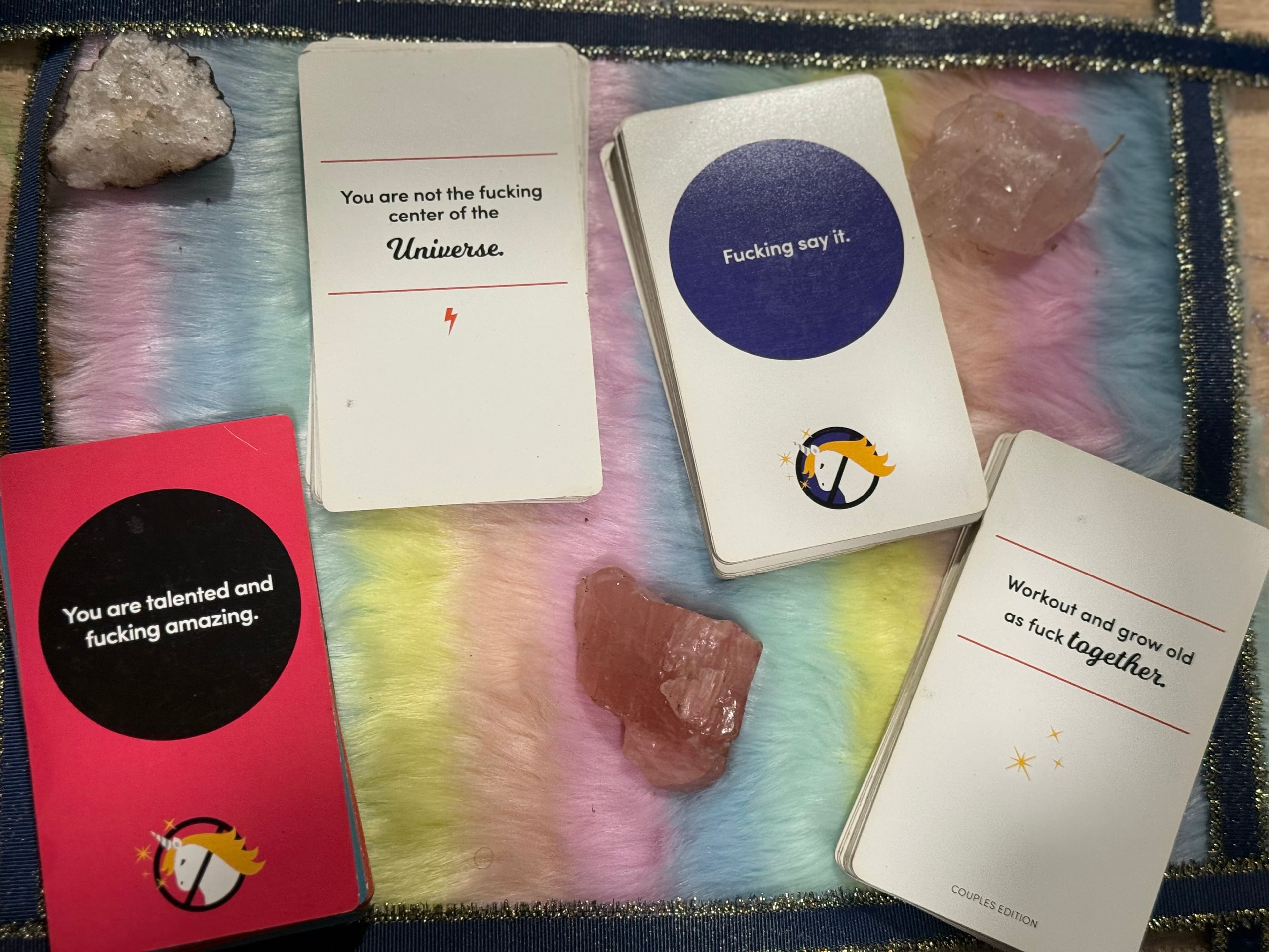 Four affirmation cards with profanity and crystals on a pastel rainbow faux fur background.