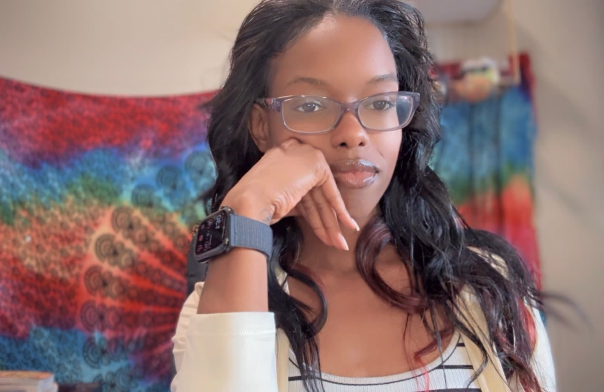 Woman wearing glasses and a smartwatch posing against a vibrant, multi-colored mandala tapestry.